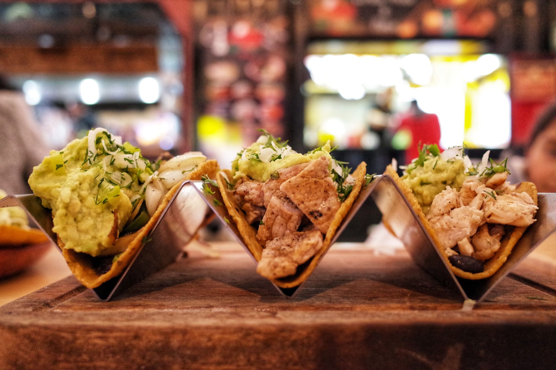 Close-up of three chicken tacos with guacamole, herbs, and onions in a restaurant setting.